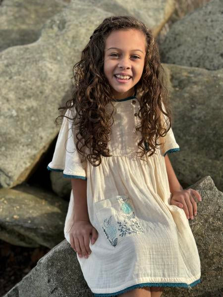 Knee length ivory dress in gauze with offset buttons on the bib, butterfly sleeves and blue trim. With floral embroidery on the patch pocket in blue and turquoise.